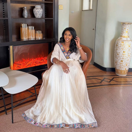 A woman wearing a white traditional Ethiopian Dress  with colorful embroidered details sits in a brown leather chair in a modern indoor setting with a decorative fireplace and large vases.