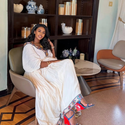 Woman in a white Ethiopian Dress  sitting in a modern room with a bookshelf and chairs.