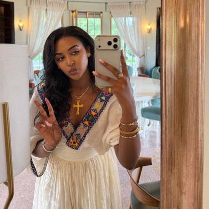 A woman taking a mirror selfie in a bright, modern room, wearing a cream-colored Ethiopian Dress  with a colorful embroidered V-neckline and gold jewelry.