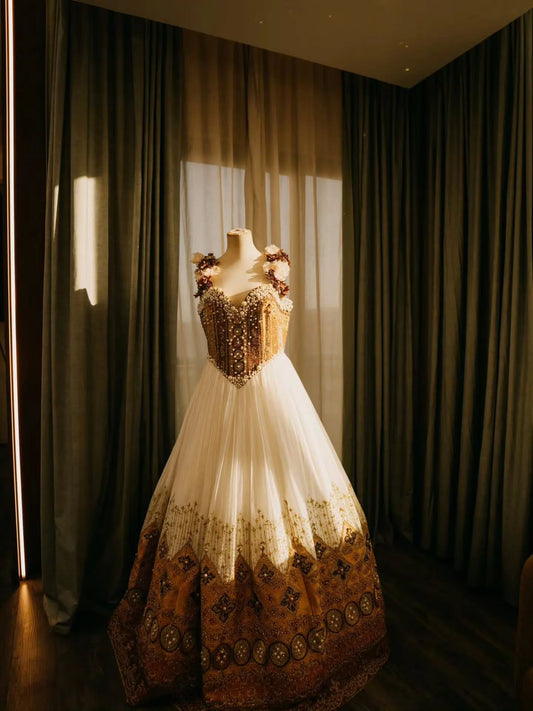 A formal ball Ethiopian dress displayed on a mannequin in a room with dark curtains and warm sunlight streaming through a window. The dress has a cream tulle skirt with ornate gold and brown embroidery concentrated on the bodice and lower skirt, and matching floral embellishments on the off-the-shoulder straps.