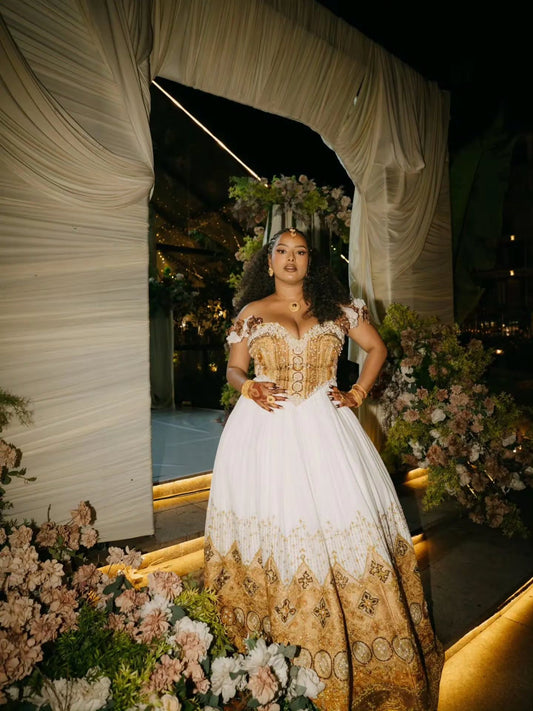 A woman wearing a formal off-the-shoulder ball Ethiopian dress with a white bodice and gold patterned skirt, standing in front of a draped event backdrop with flowers and warm lighting.