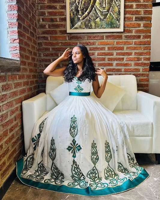 Woman in a white and green Habesha Dress sitting on a white chair against a brick wall.