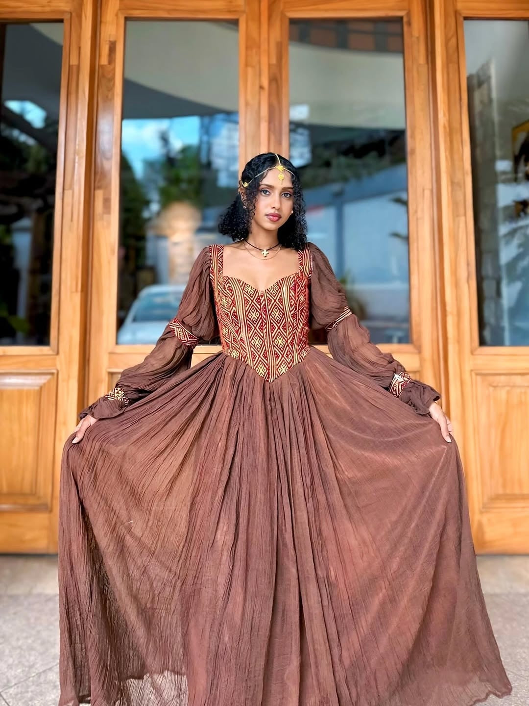 Woman in a long brown Ethiopian traditional dress standing in front of wooden doors.