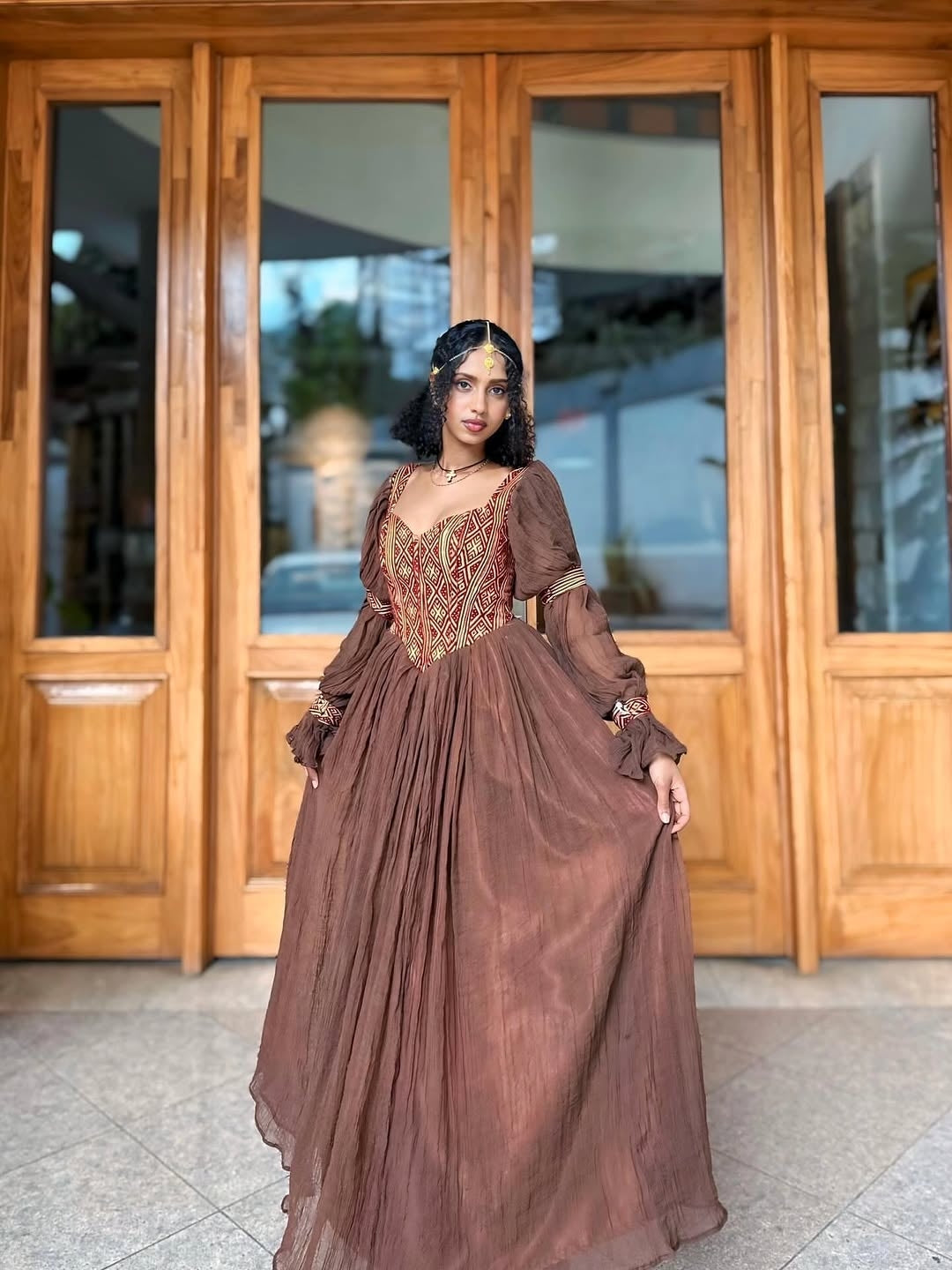 Woman in a brown Ethiopian traditional dress standing in front of wooden doors.
