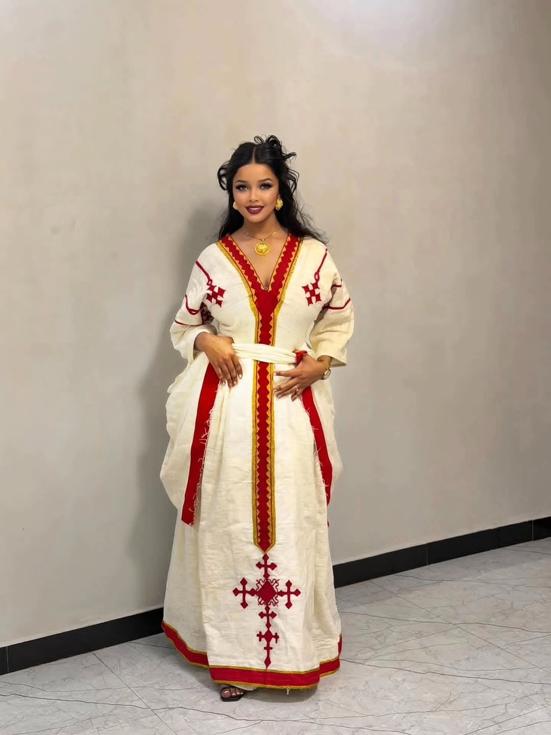 Woman wearing a white dress with red and gold Ethiopian dress embroidery against a plain background