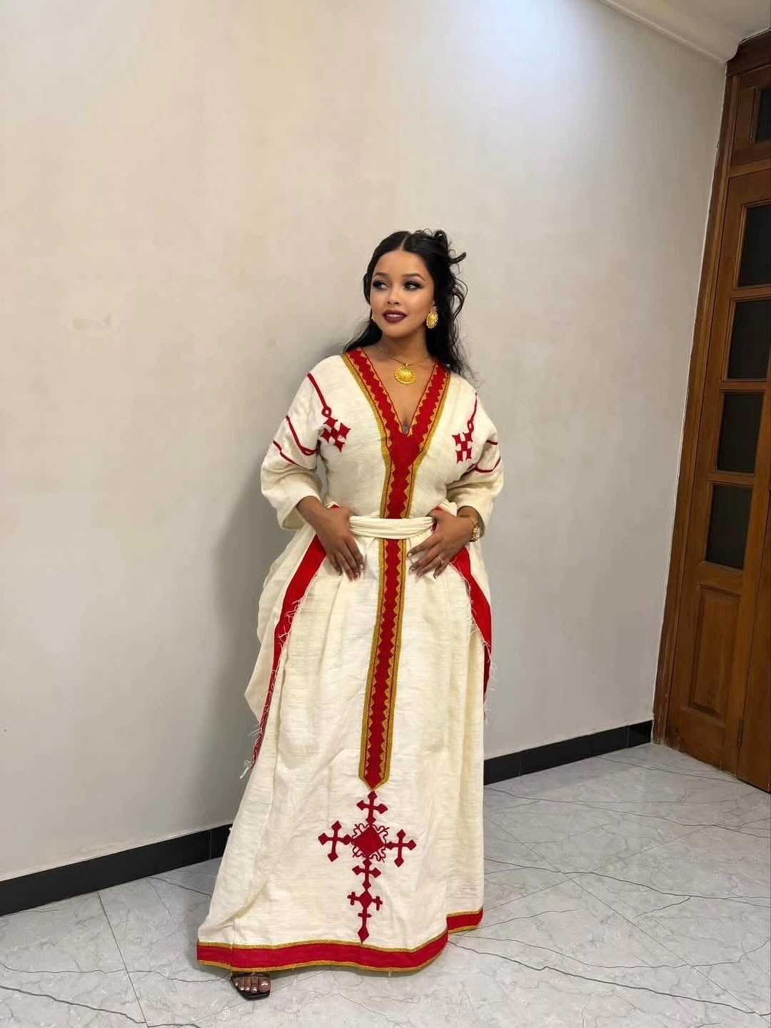 Woman wearing a white Ethiopian dress with red embroidery in a room with a wooden door.