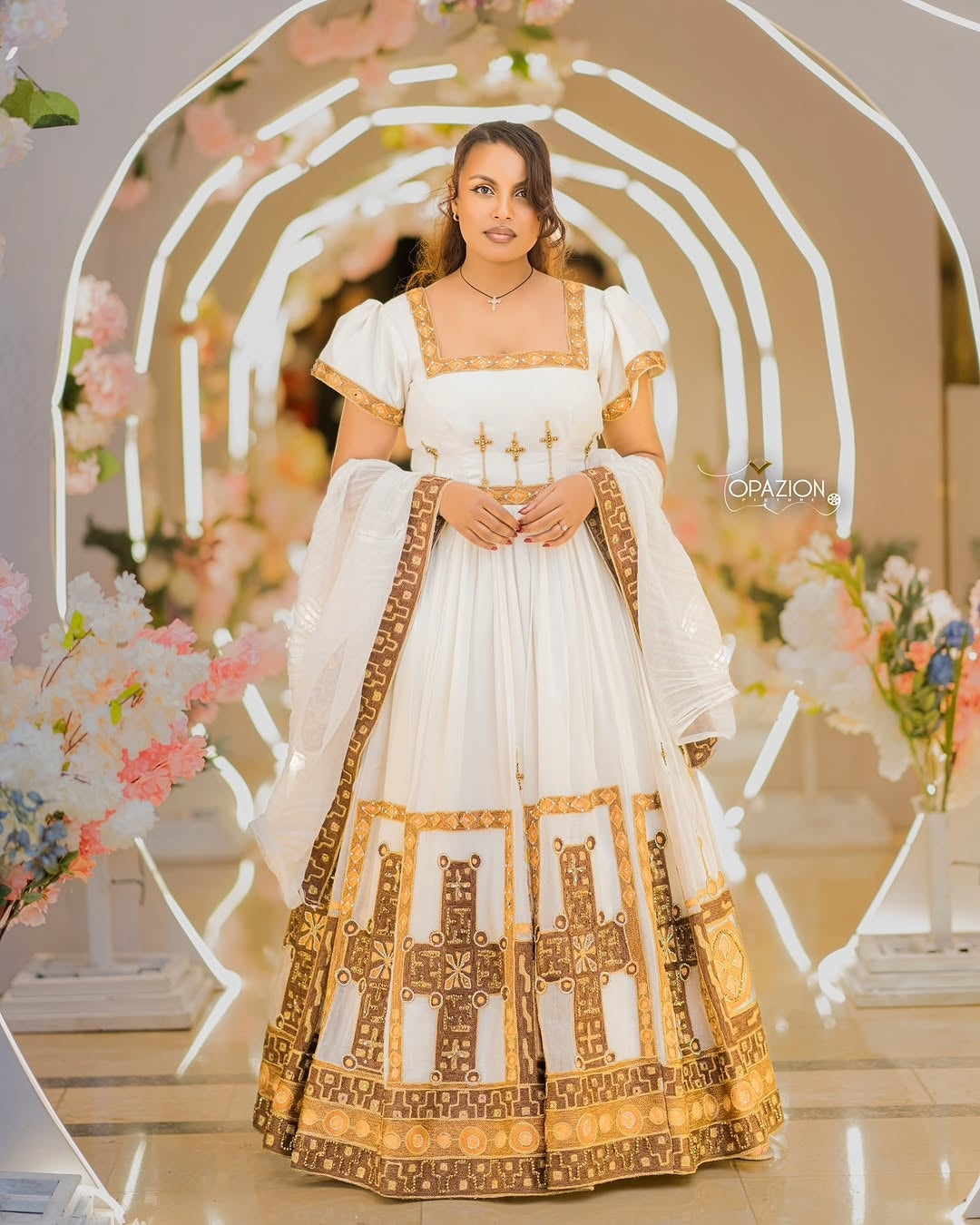 Woman in a white and gold Ethiopian Traditional Wedding Dress standing in front of floral decorations.