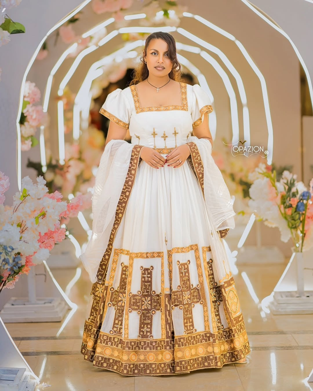 Woman in a white and gold Ethiopian Traditional Wedding Dress standing in front of a decorative arch with flowers.