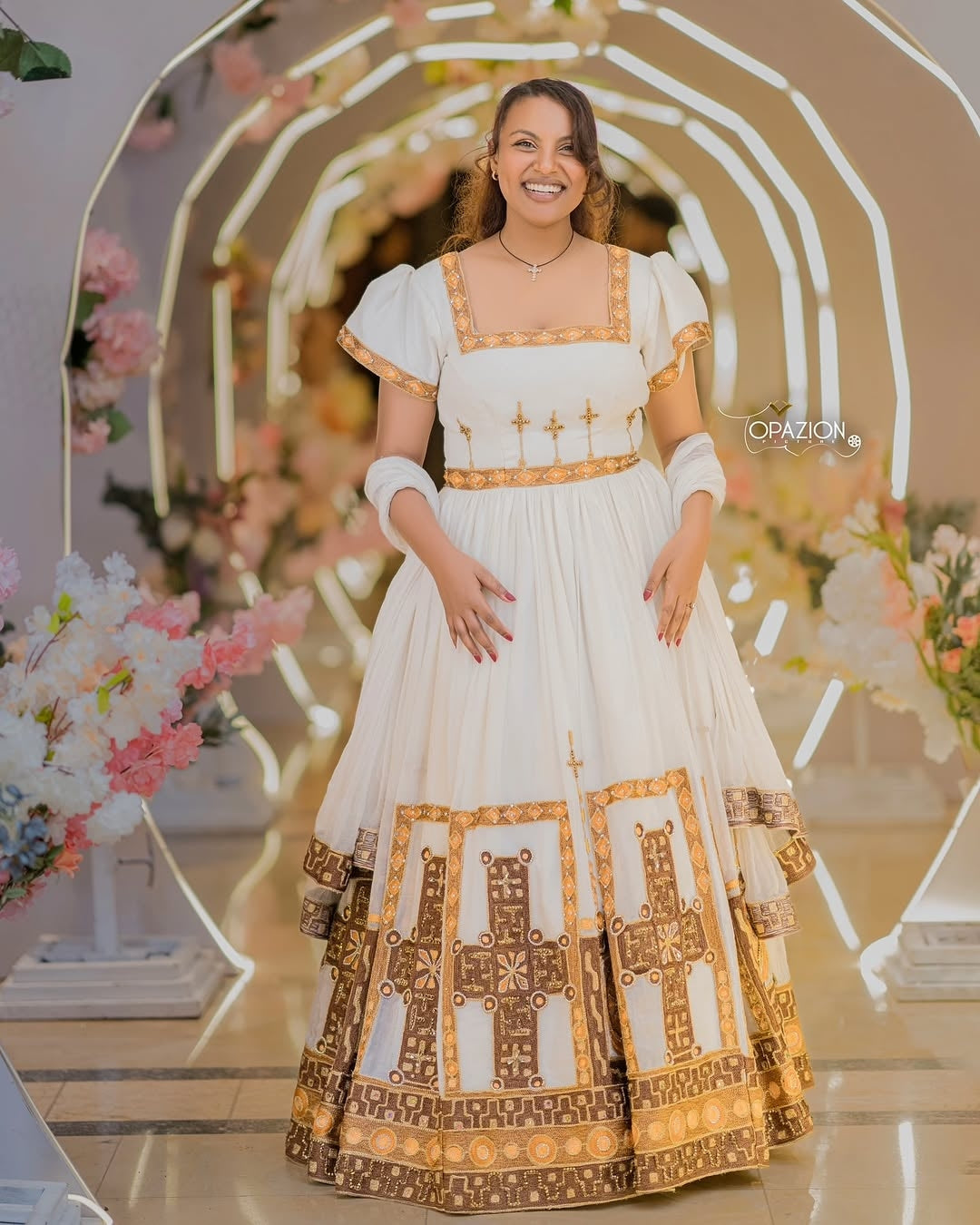 Woman in a white and gold Ethiopian Traditional Wedding Dress standing in front of floral decorations.
