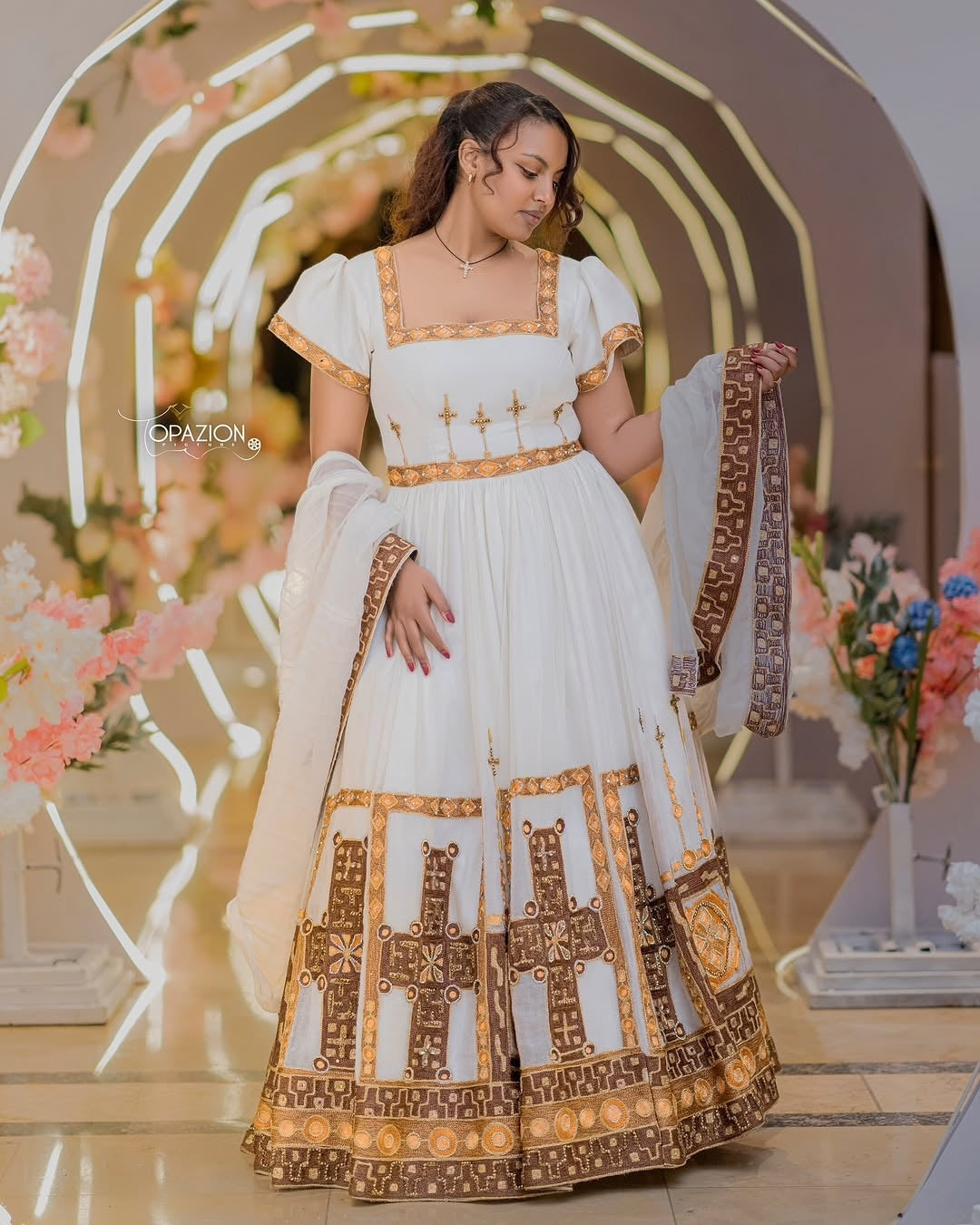 Woman in a white and gold Ethiopian Traditional Wedding Dress standing in a decorated indoor setting.