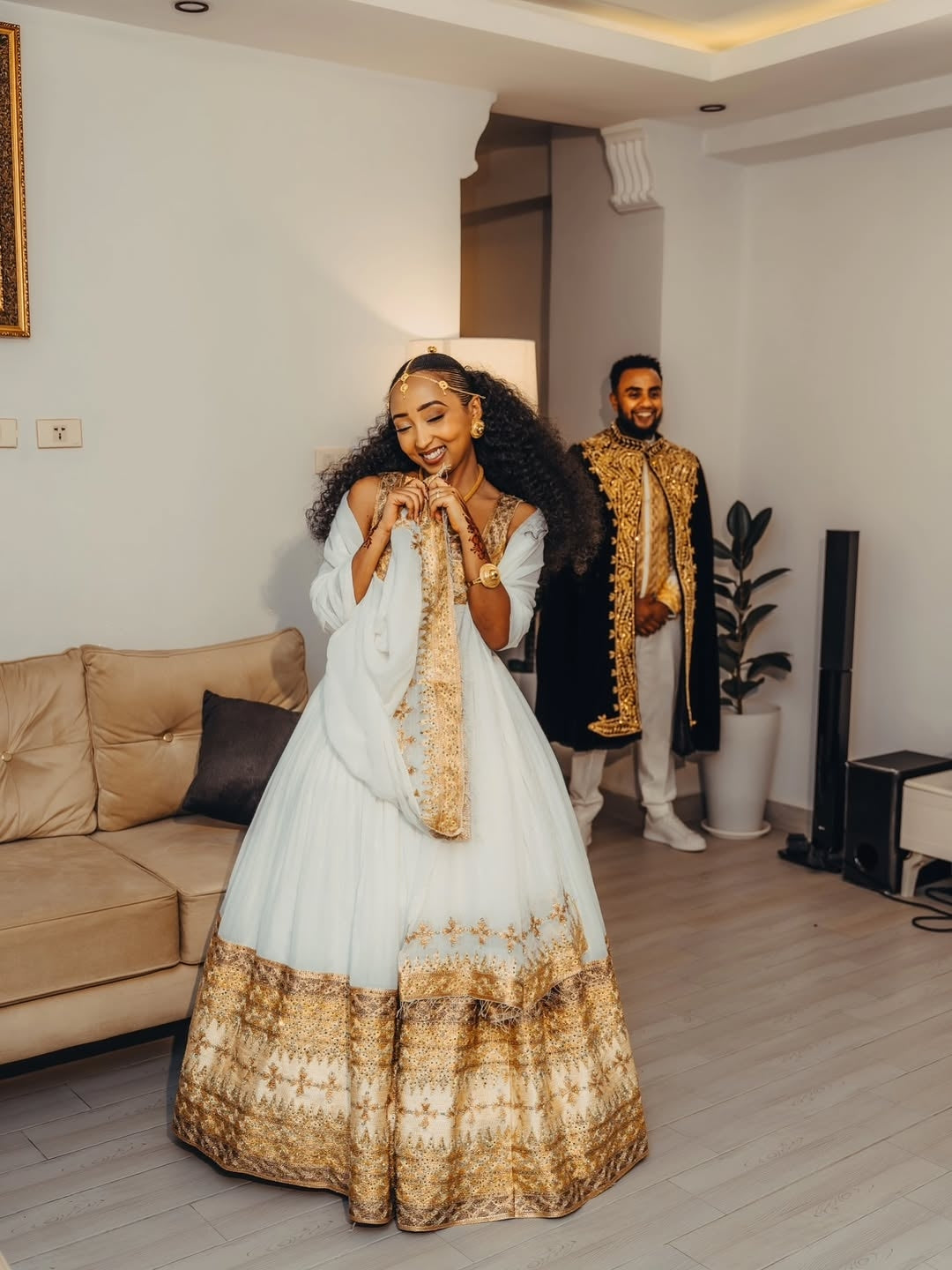 Woman in a white and goldGold Ethiopian Matching Couple Wedding Set standing in a living room with a man in formal attire in the background.