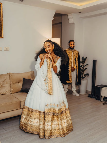 Woman in a white and goldGold Ethiopian Matching Couple Wedding Set standing in a living room with a man in formal attire in the background.