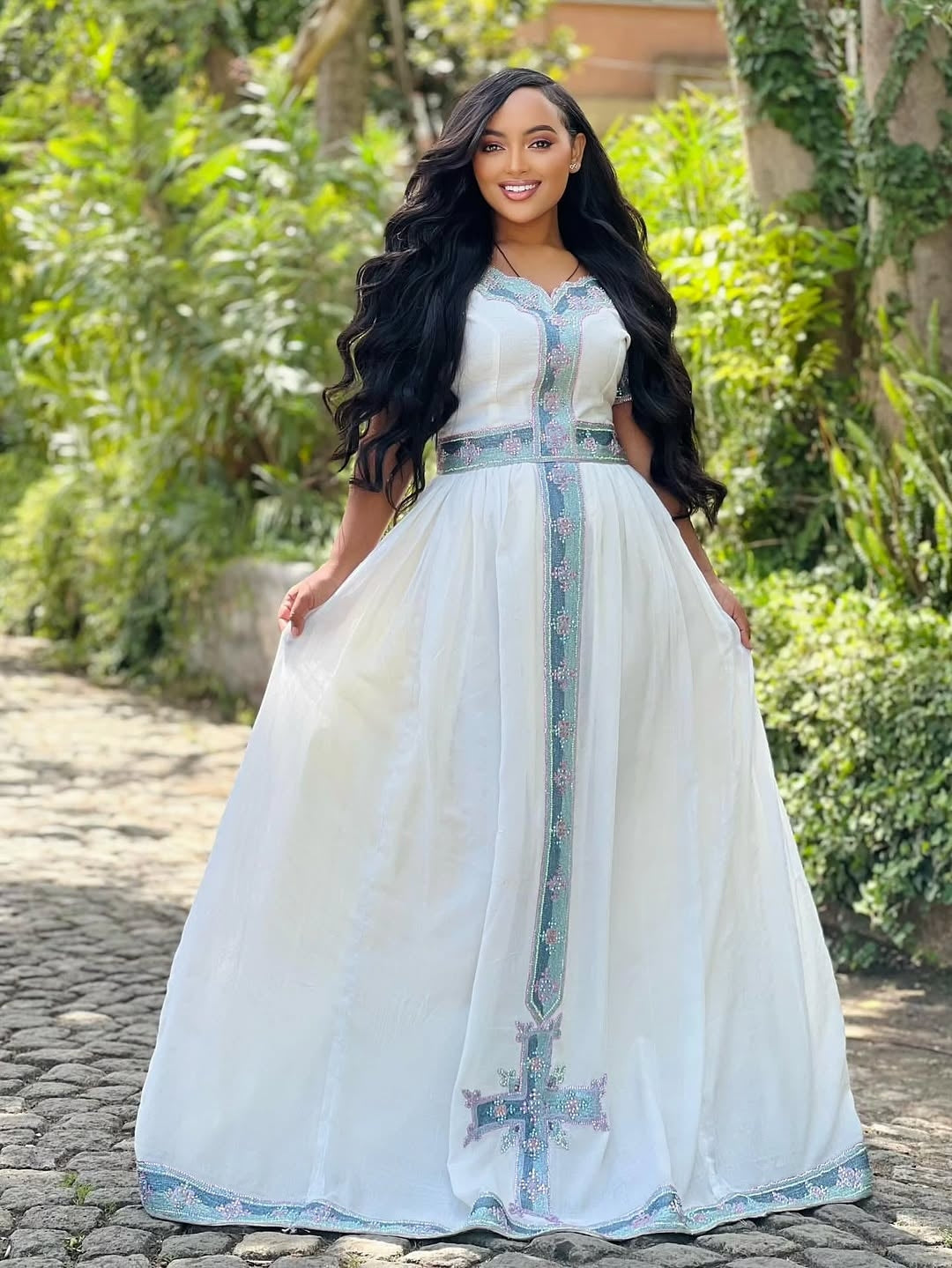 Woman in a white Ethiopian dress with floral patterns standing outdoors on a stone path.