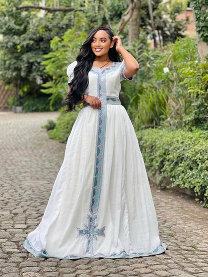 Woman in a long, light-colored dress with floral patterns standing on a stone path with greenery in the background.