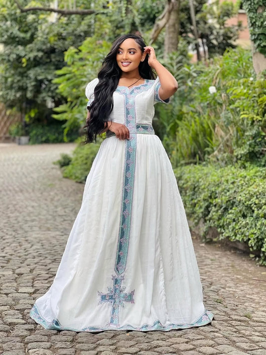 Woman in a long, light-colored dress with floral patterns standing on a stone path with greenery in the background.