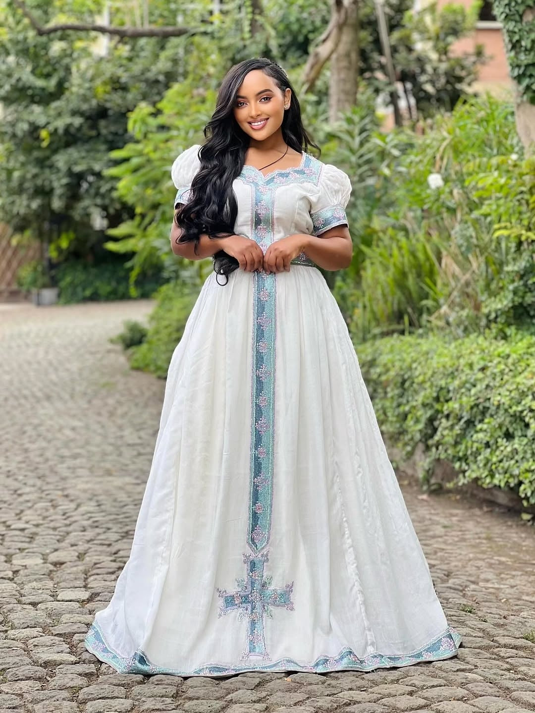 Woman in a white dress with blue embroidery standing outdoors on a stone path.