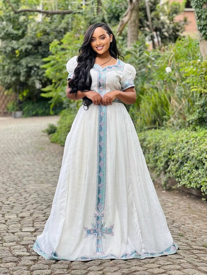 Woman in a white dress with blue embroidery standing outdoors on a stone path.