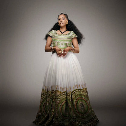 A woman stands Habesha Wedding Dress  a plain gray studio background wearing a long white gown with ornate green and gold patterned detailing on the bodice and hem.