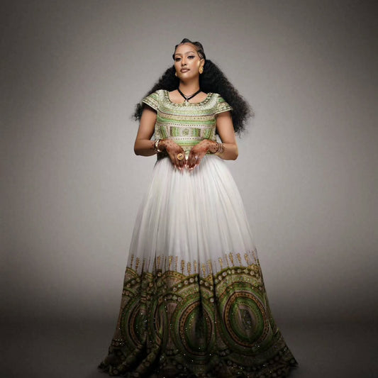 A woman stands Habesha Wedding Dress  a plain gray studio background wearing a long white gown with ornate green and gold patterned detailing on the bodice and hem.