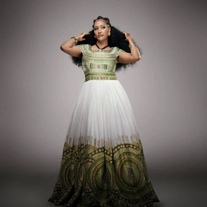 A woman modeling a floor-length white Habesha Wedding Dress  with green and gold ornate embroidery concentrated on the bodice and lower hem, photographed against a plain gray studio background.