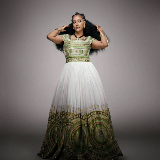 A woman modeling a floor-length white Habesha Wedding Dress  with green and gold ornate embroidery concentrated on the bodice and lower hem, photographed against a plain gray studio background.