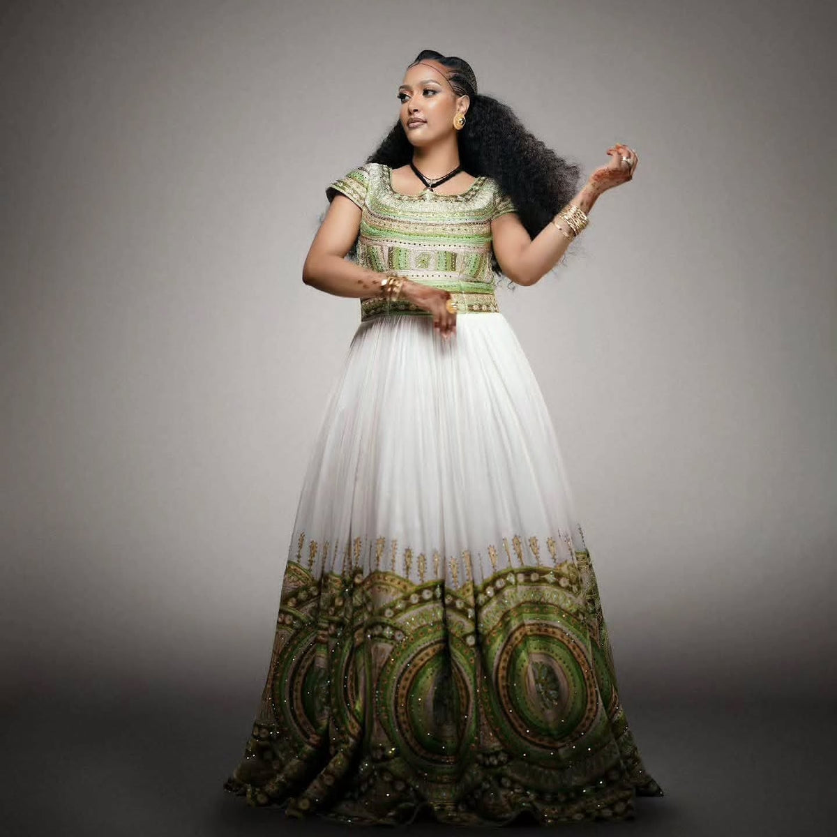 A woman modeling a traditional African Habesha Wedding Dress  with a green and white patterned bodice and a voluminous white skirt featuring a green and gold circular pattern at the hem, set against a dark gray studio background.