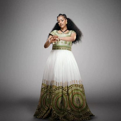 A woman modeling a traditional long Habesha Wedding Dress  with a white tulle skirt and an ornate green, gold, and red patterned bodice and hem, posed against a solid gray studio background.