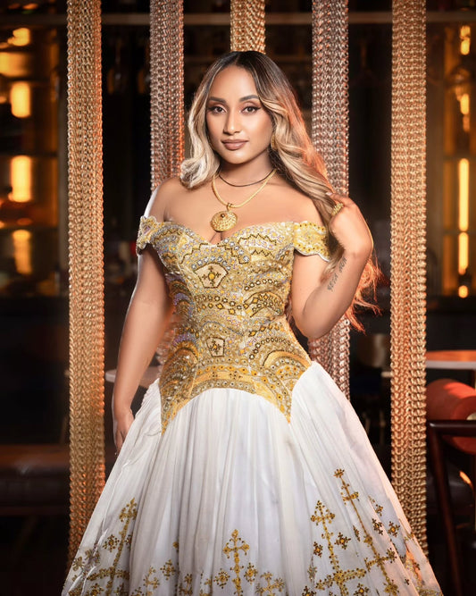 A woman modeling an off-the-shoulder white Ethiopian Dress for Women with ornate gold embroidery and embellishments, standing in front of a decorative gold chain backdrop.