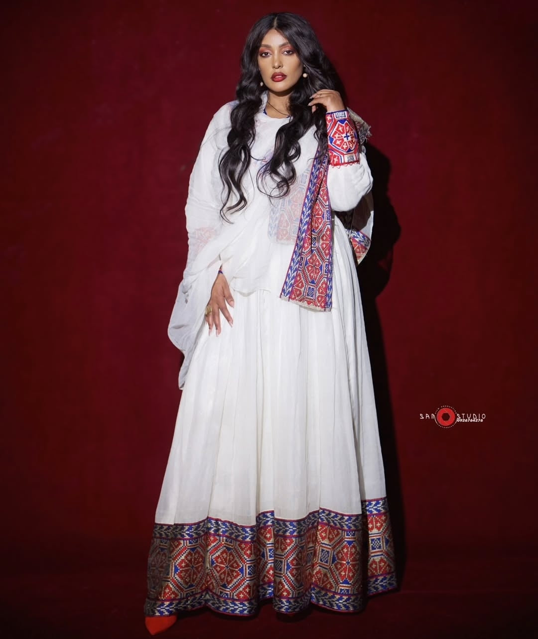 Woman wearing a white Ethiopian Habesha Dress with colorful patterns against a red background
