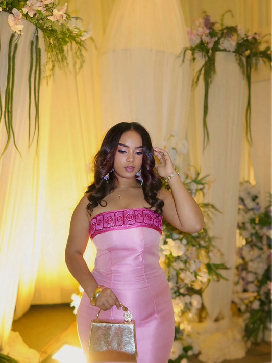 A woman wearing a pink strapless satin Habesha kemis  with a floral embroidered band across the chest, holding a metallic silver clutch bag, posed in front of a warm yellow draped backdrop decorated with flowers.