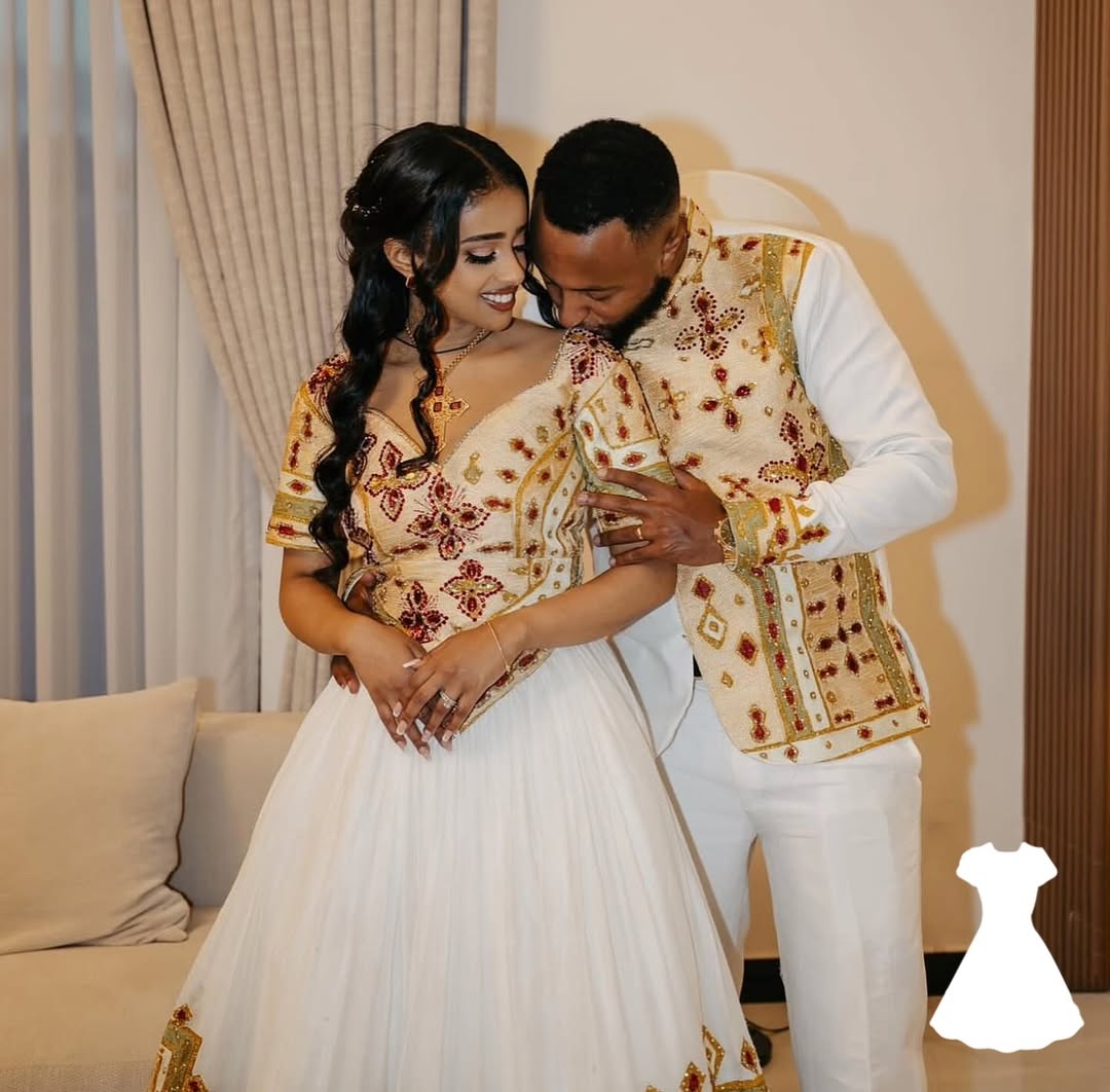 Couple in traditional wedding attire embracing in a room with beige curtains and a couch.