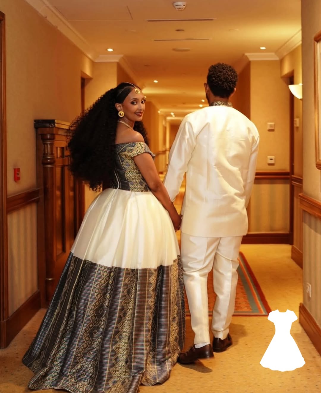 Woman in a Ethiopian traditional wedding dress holding hands with a man in a white outfit in a hallway.