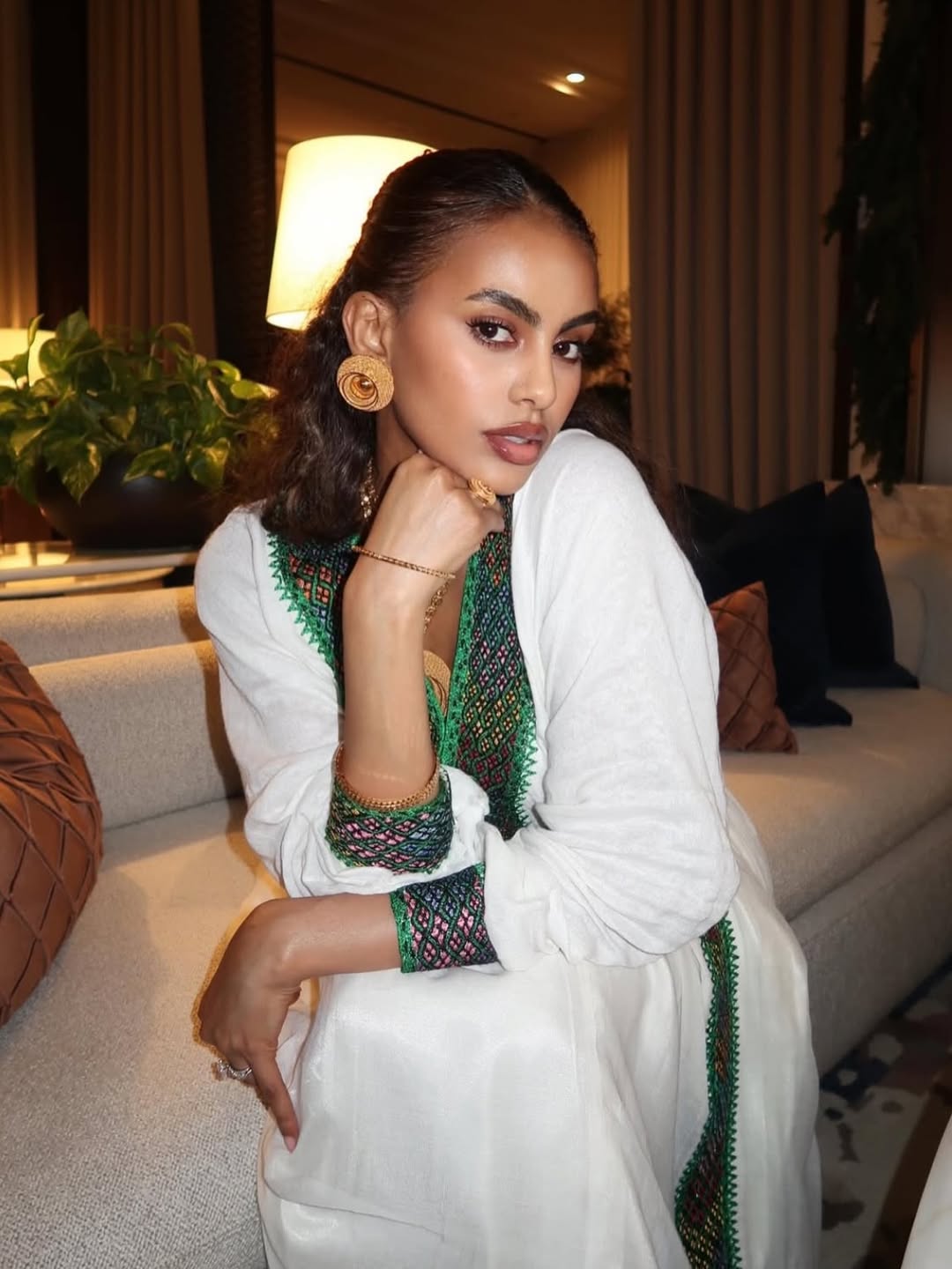 Woman in a white and green traditional Ethiopian traditional dress sitting on a couch in a well-lit room.