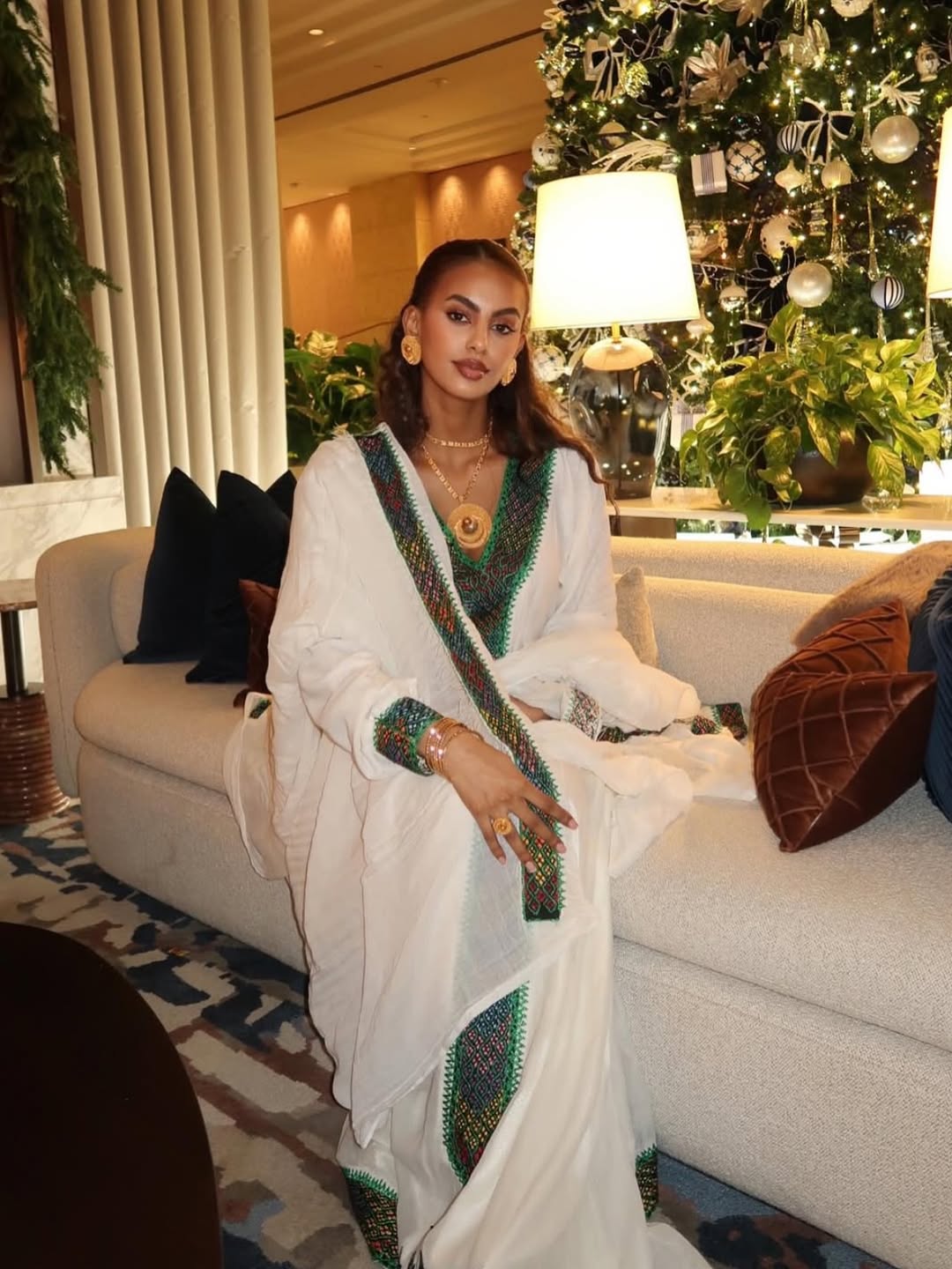 Woman in a white Ethiopian traditional dress  with green and gold patterns sitting on a couch in a decorated room.