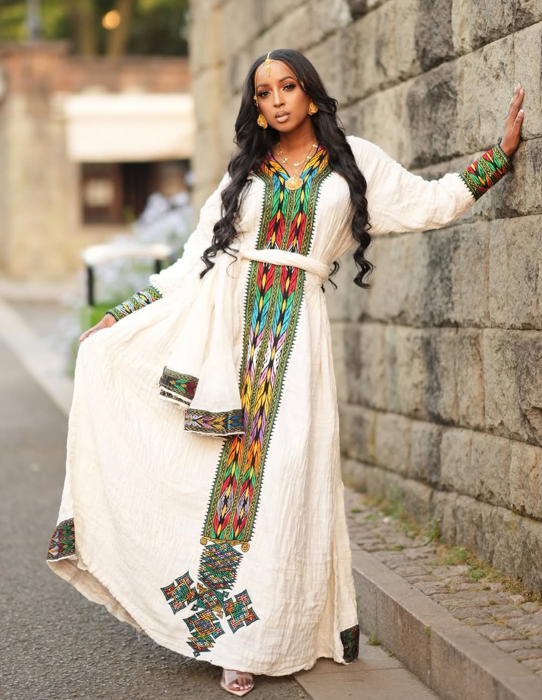 Woman in a long, light-colored Ethiopian traditional dress with a colorful scarf standing against a stone wall.