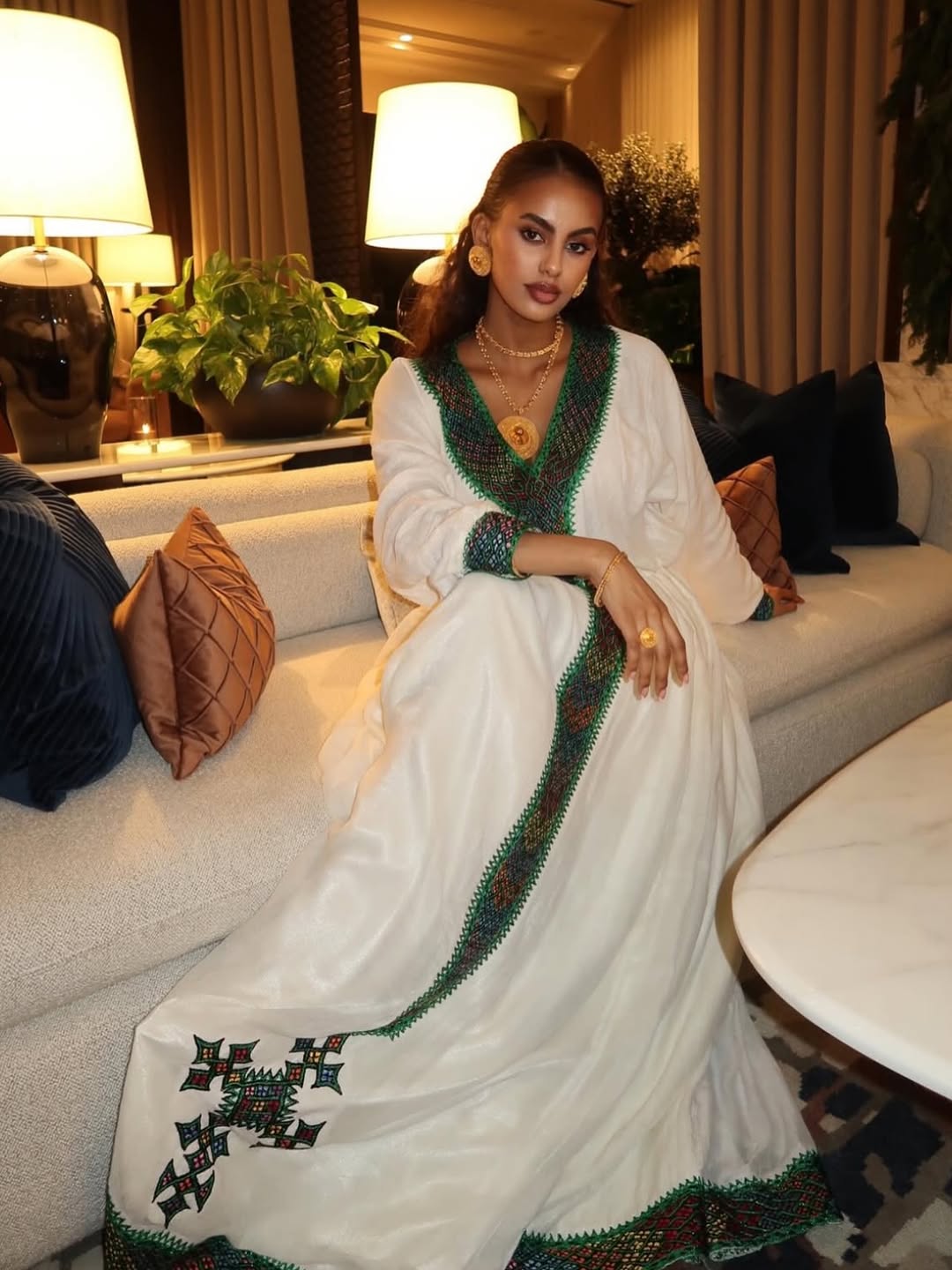 Woman in a white Ethiopian traditional dress  with green and black embroidery sitting on a couch in a stylish room.