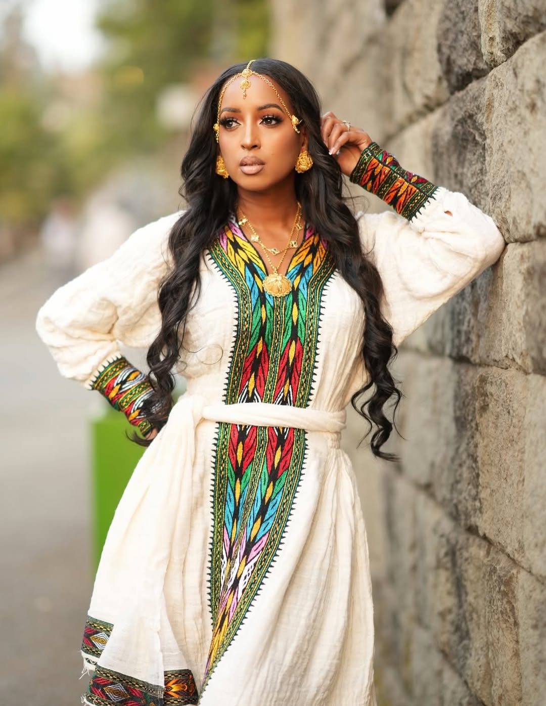 Woman in a Ethiopian traditional outfit with colorful patterns leaning against a stone wall.