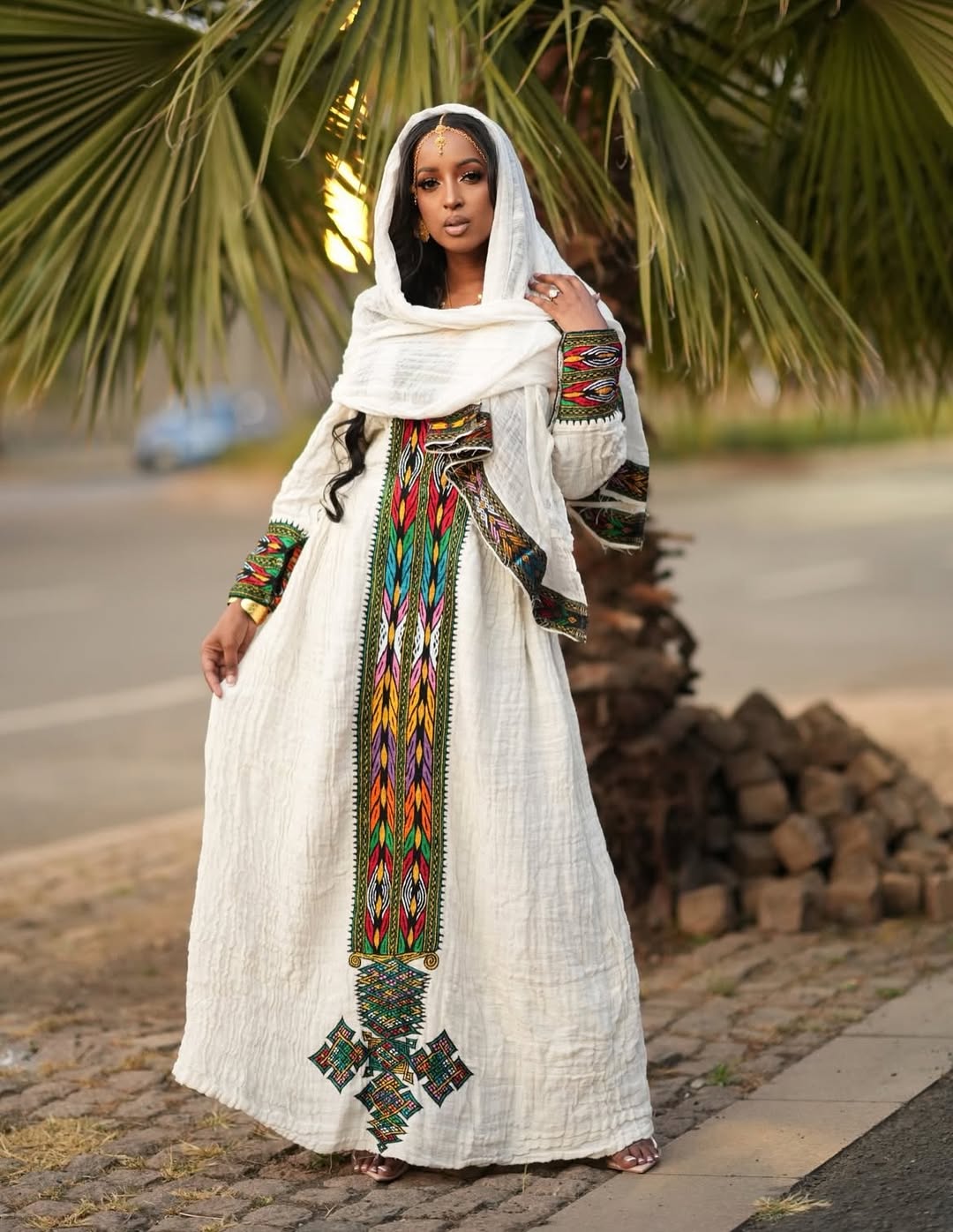 Woman in a long, embroidered white Ethiopian traditional dress with colorful patterns standing outdoors.