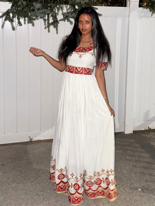 A woman wearing a white maxi Habesha Zuria  with red and gold bohemian-style embroidery, standing in front of a white fence.