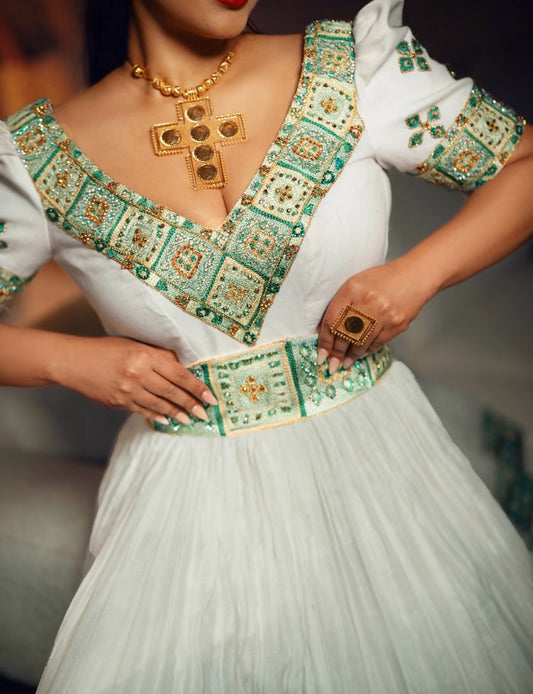 A close-up of a woman wearing a white traditional Ethiopian dress for women with ornate green and gold embroidered patterns along the neckline, sleeves, and waist. She is also wearing a gold cross necklace and a gold ring.