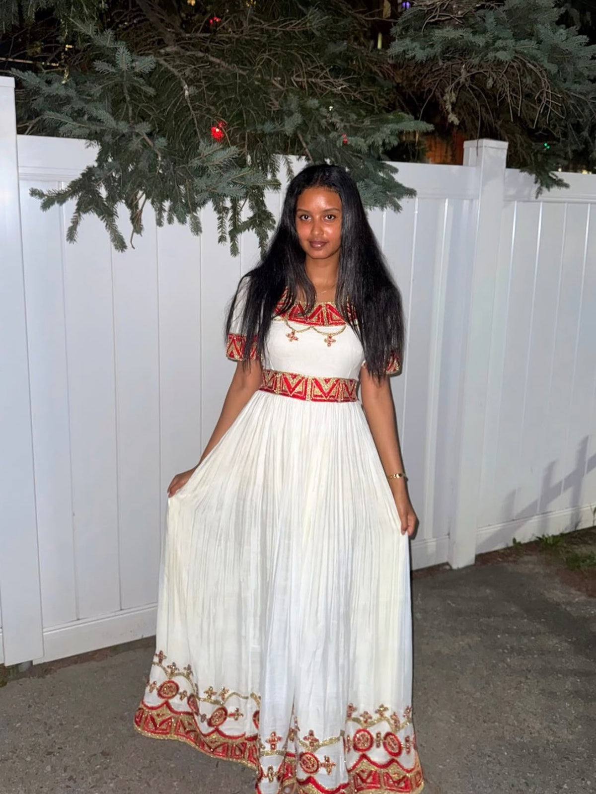 A woman wearing a long white maxi Habesha Zuria  with red and gold geometric embroidery, standing in front of a white fence at night.