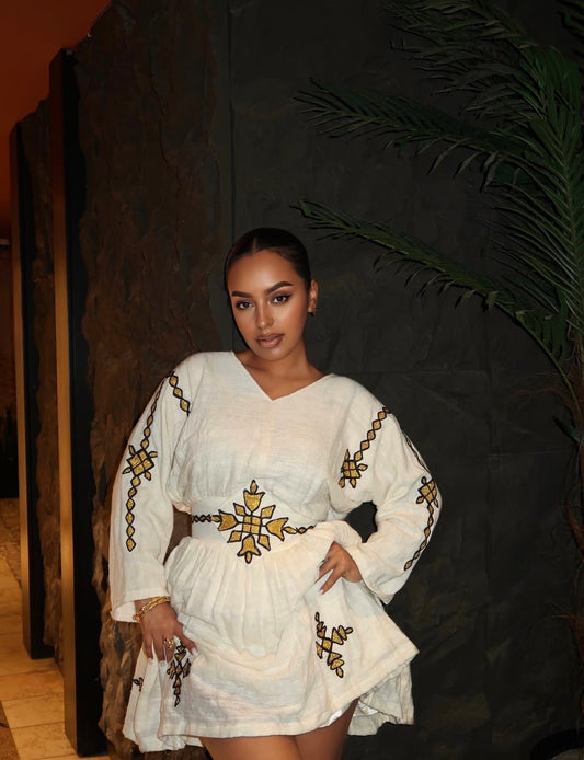 A woman modeling a white long-sleeve Ethiopia Clothes  with gold and black traditional embroidery, standing against a dark textured wall with a plant in the background.