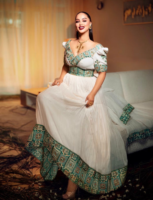 A woman models a white off-the-shoulder maxi Ethiopian dress for women with intricate teal and gold patterned embroidery on the bodice, waist, hem, and sleeve cuffs, posed in a warmly lit indoor setting.