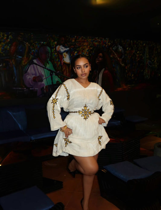 A woman posing in a white traditional-style Ethiopia Clothes  with gold embroidery, set against a dark, decorated indoor background.