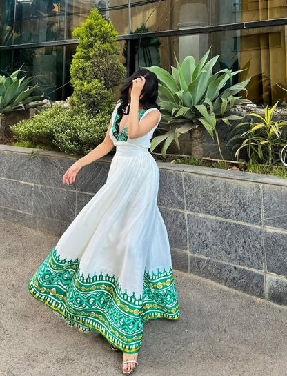 Woman in a white Ethiopia traditional dress with green patterns walking outdoors near a building with plants.