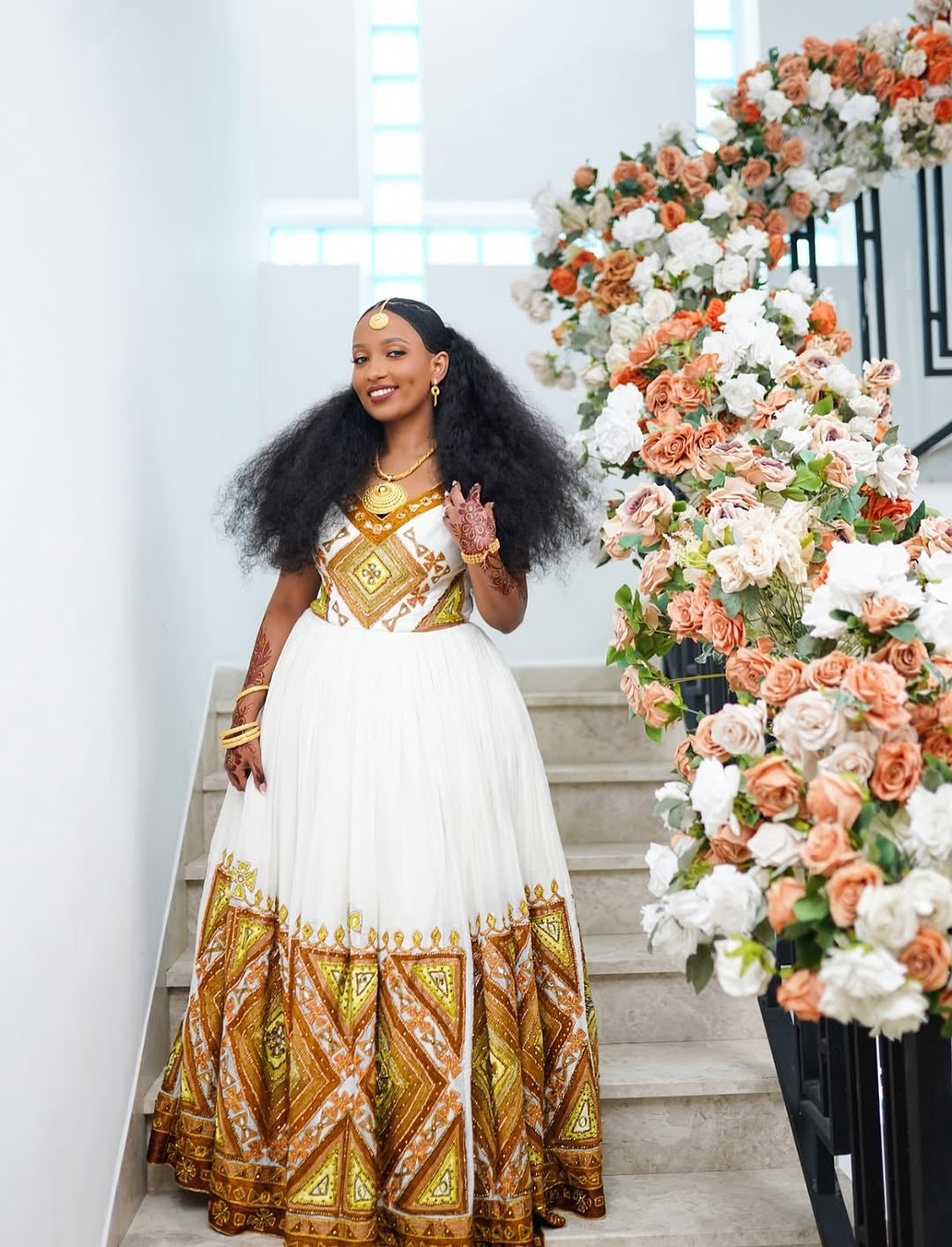 Woman in a white and gold Ethiopia traditional wedding dress standing next to a floral arch.