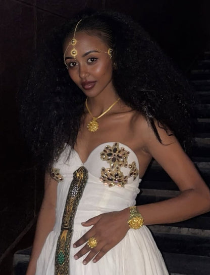 Woman in a white Ethiopian traditional dress with gold embellishments and jewelry against a dark background