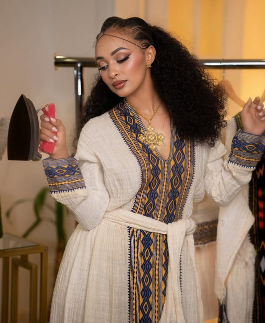 Woman in a patterned Eritrean Dress
 holding an iron in a room with a blurred background