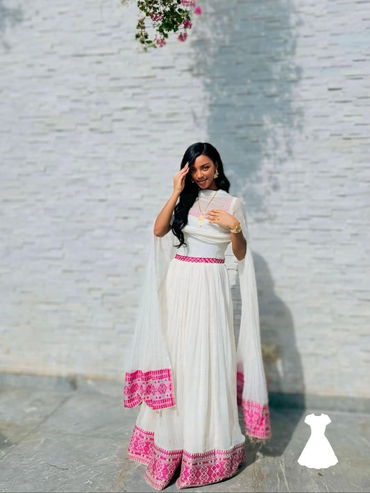 Woman wearing a white and pink traditional Ethiopian Kemis against a light gray wall.