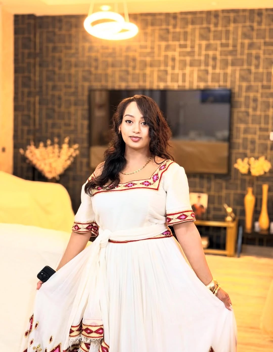 Woman in a white Ethiopian Libs with red and gold patterns standing in a room with decorative elements.